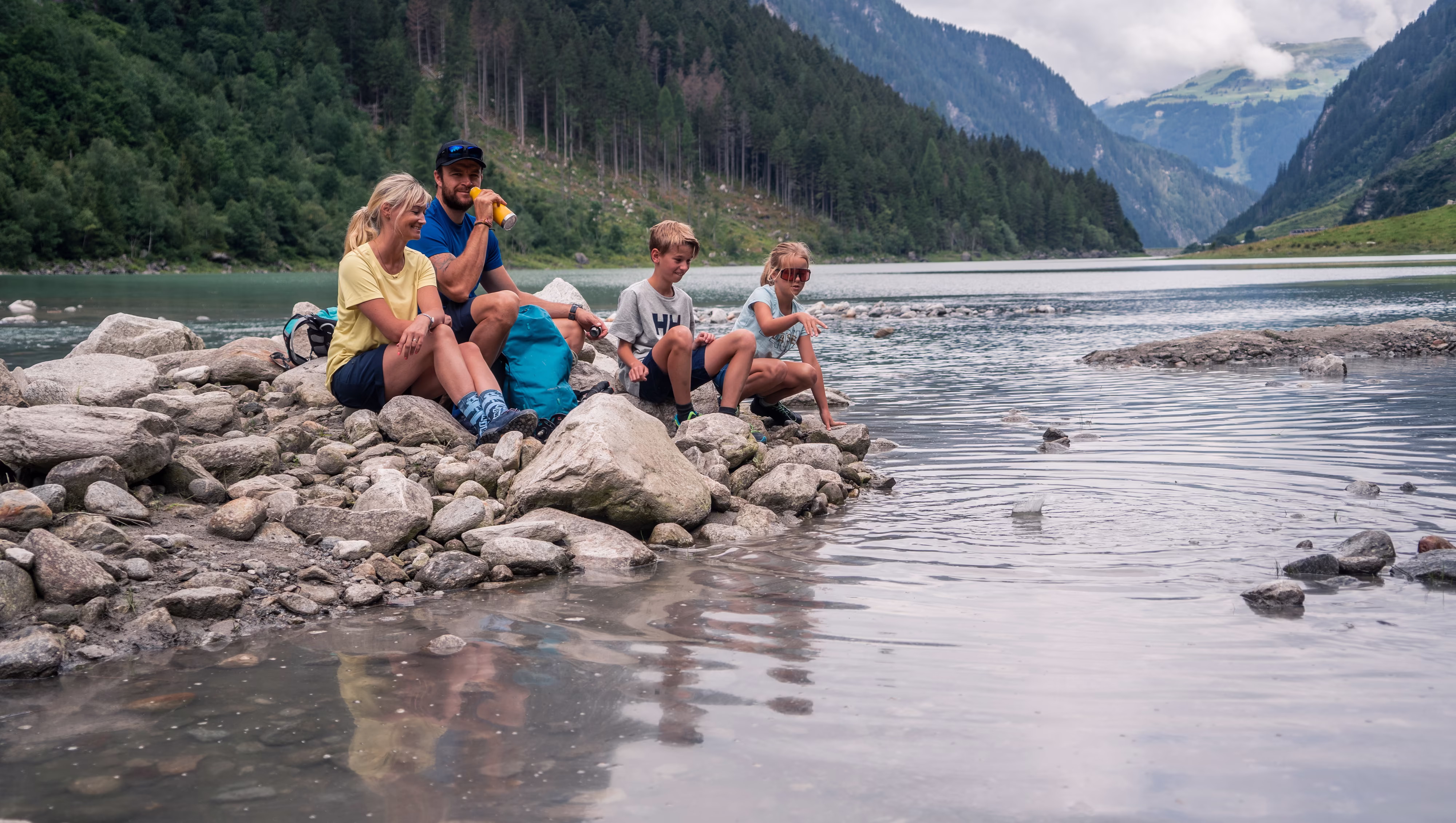 Familie sitzt an einem Bergsee auf Steinen und jausnet 